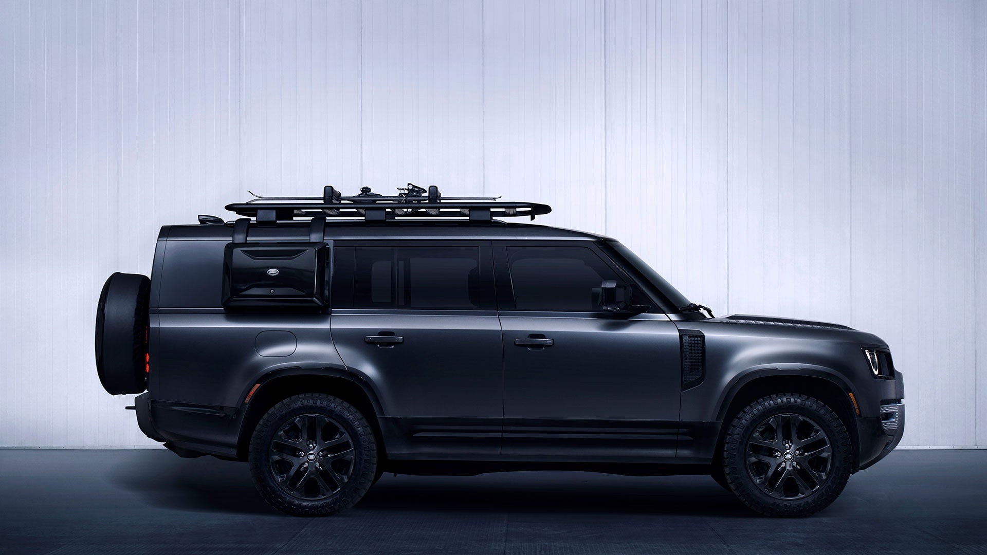 A dark gray Range Rover Defender parked in front of white wall with roof rack on top